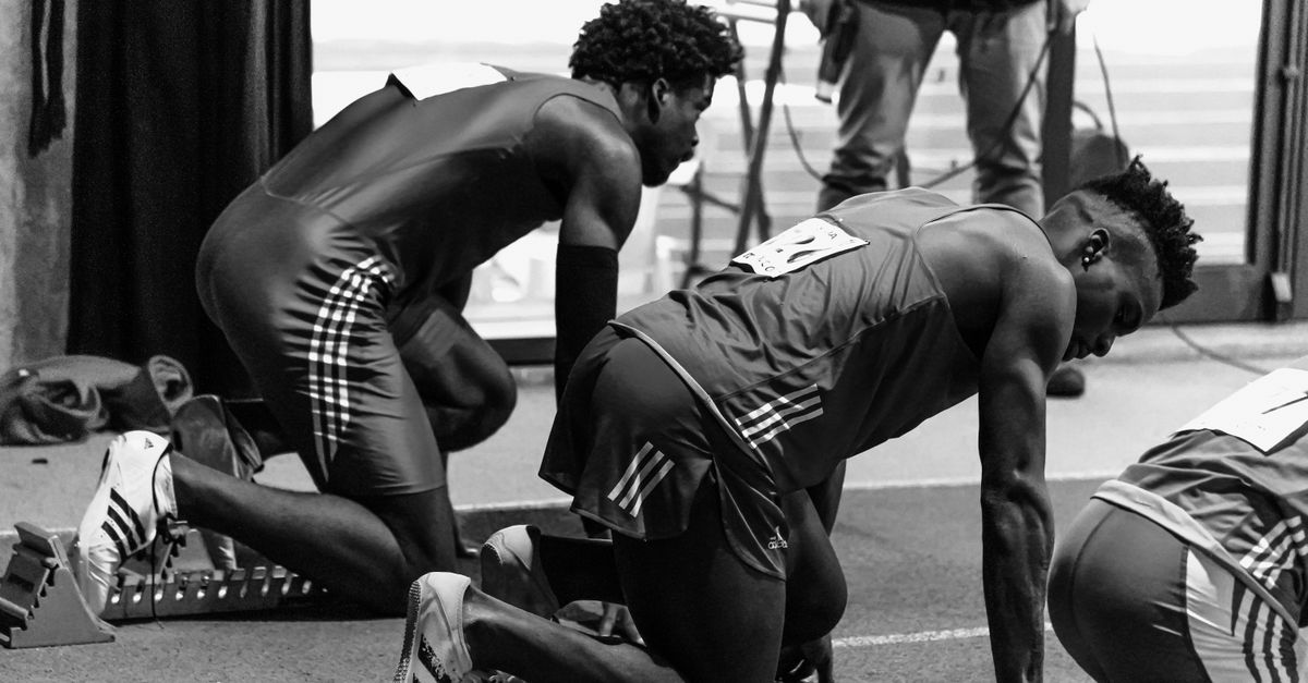 Black and white image of athletes in starting blocks ready for a sprint race indoors.